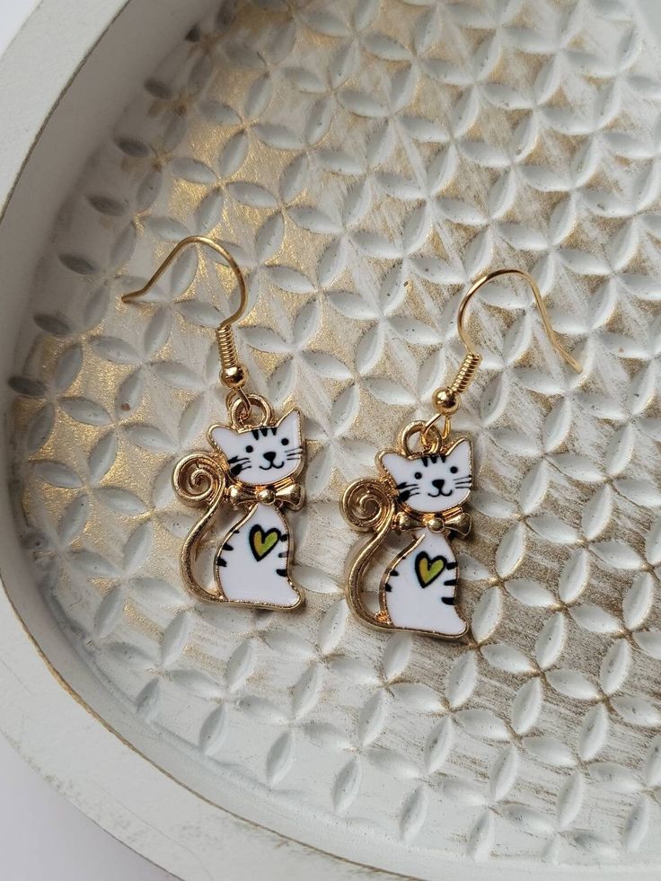 These cute, white cat earrings are so sweet! The little kitty even has a heart on its chest, making it even more endearing! The drop length is about 1.5 inches and the width is about .5 inches. They are made of zinc alloy and enamel. These whimsical earrings are lightweight making them comfortable to wear and ideal for older kids, teens, and adults. The ear wires are gold in color, hypoallergenic, and nickel-free. Each pair comes with two, clear rubber backs. They would make a perfect gift for a Gold Cat Design Earrings For Gifts, Gold Metal Cat Design Earrings, Gold Metal Earrings With Cat Design, White Heart Earrings With Ear Wire As A Gift, Cute Metal Jewelry With Cat Design, Cute Cat Print Jewelry Gift, White Metal Heart Earrings For Gift, White Metal Heart Earrings Gift, Elegant White Cat Design Jewelry