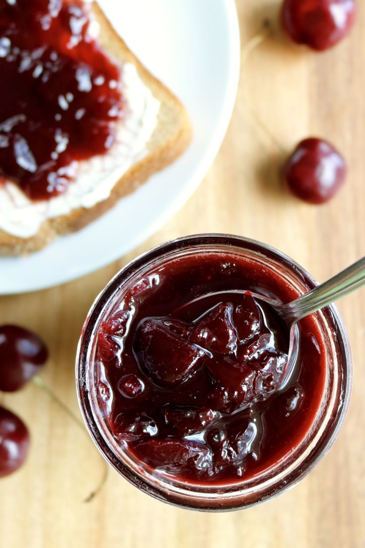 a glass jar filled with jelly next to bread and cherries on a wooden table