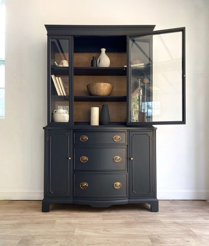 a black china cabinet with glass doors and drawers on the bottom, in front of a white wall