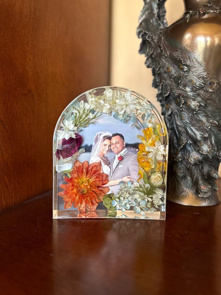 a glass photo frame sitting on top of a wooden table next to a vase with flowers