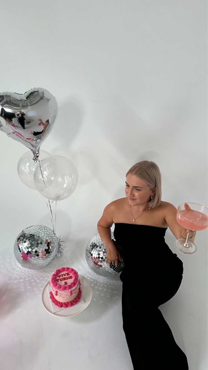 a woman in a black dress sitting next to a pink cake and some silver balloons