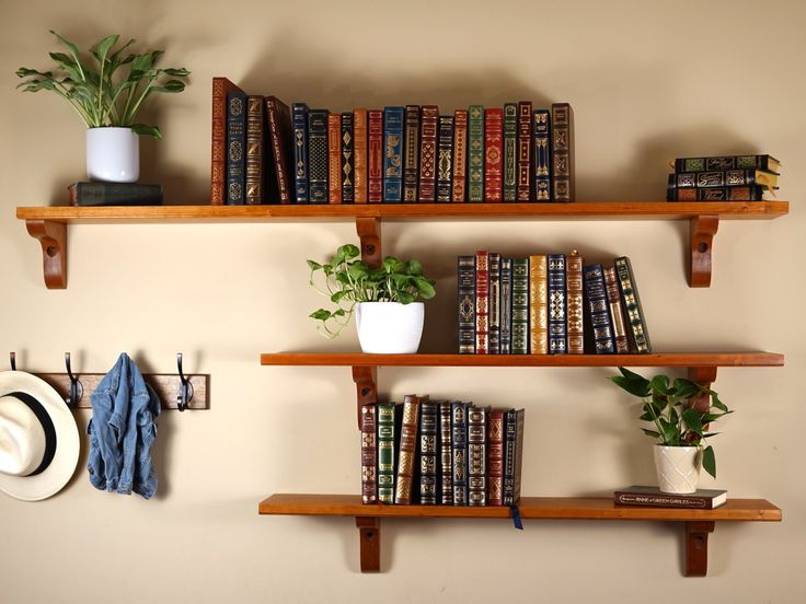 three wooden shelves with books, hats and plants on them against a wall filled with books