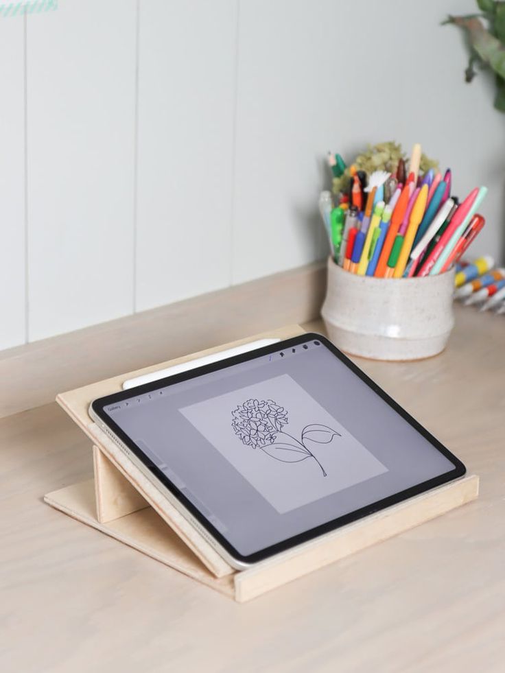 an ipad on a table with colored pencils in the background and a potted plant next to it