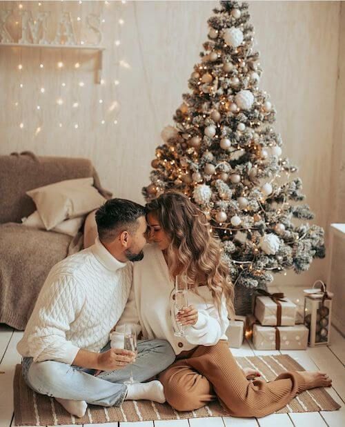 a man and woman sitting on the floor next to a christmas tree holding wine glasses