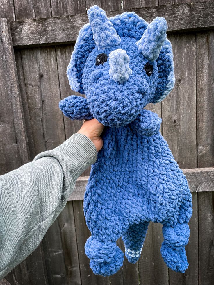 a hand holding a blue knitted elephant toy in front of a wooden fence background