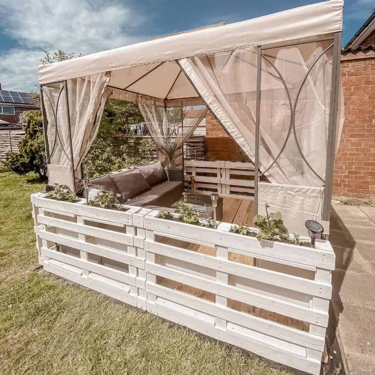 an outdoor living area with wooden pallets and white curtains on the side of it