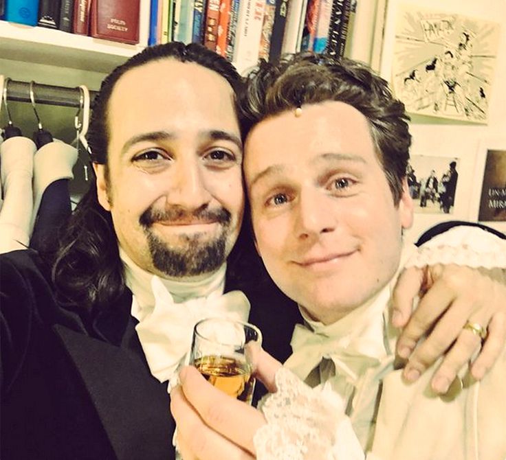 two men in tuxedos pose for a photo with their wine glasses and books behind them