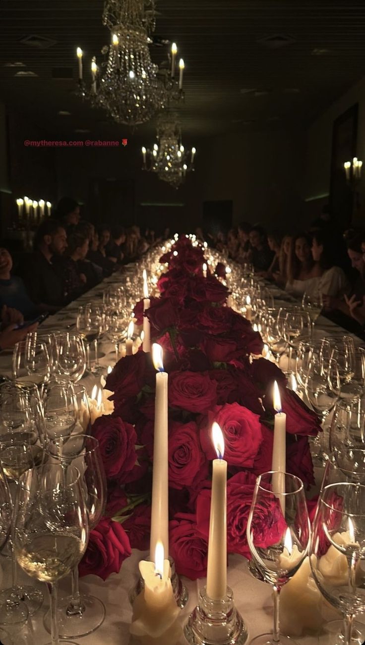 a long table with many candles and wine glasses on it, surrounded by red roses