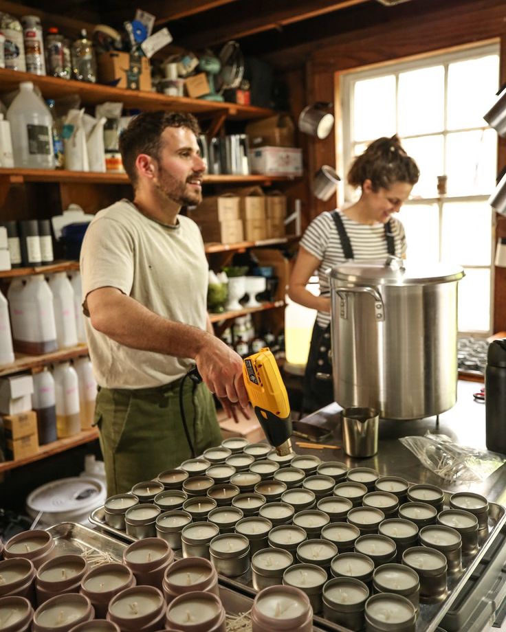 two people are working in a kitchen with many jars on the counter and one person is pouring something into a pot