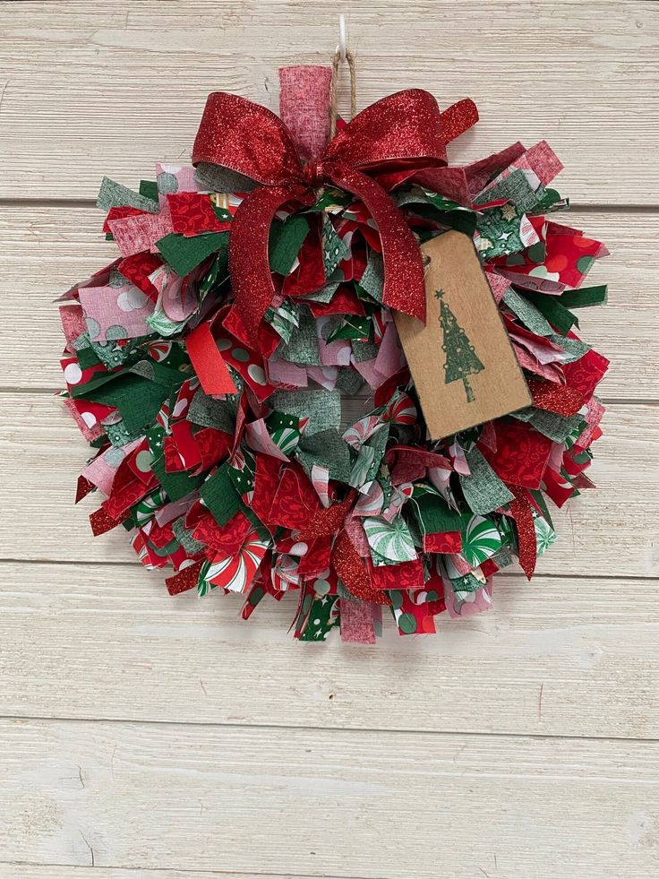 a red and green christmas wreath with a tag on it hanging on a wooden wall