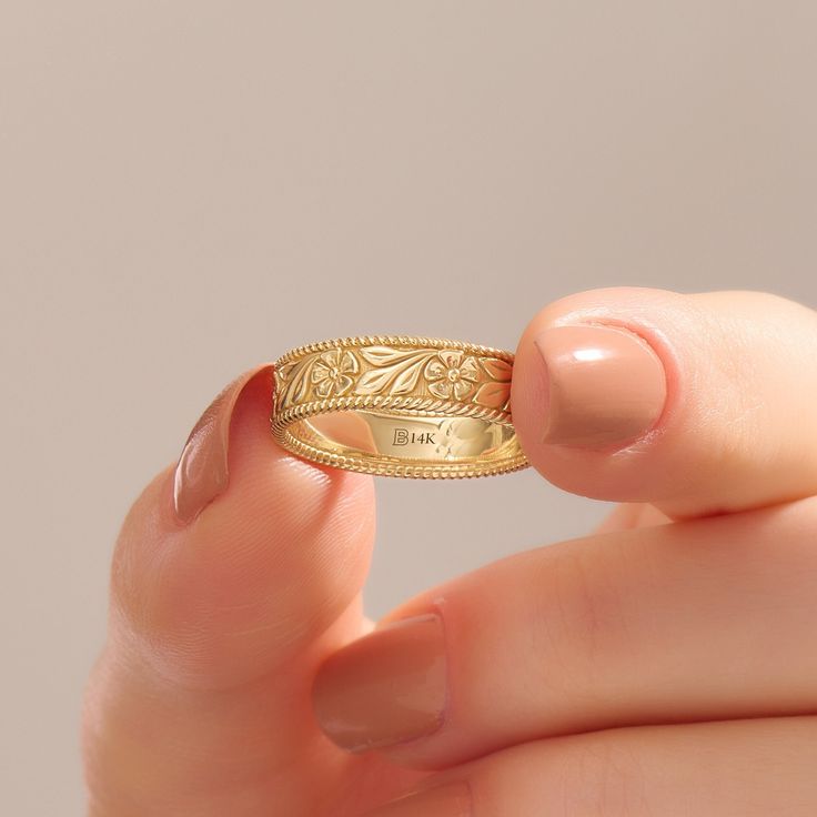 a woman's hand holding a gold ring with leaves on the band and engraving