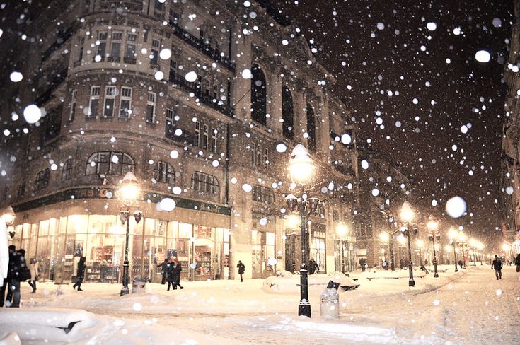 people are walking in the snow on a city street at night with lights and buildings