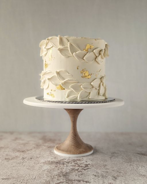 a white cake with gold leaf decorations on a wooden stand in front of a gray wall