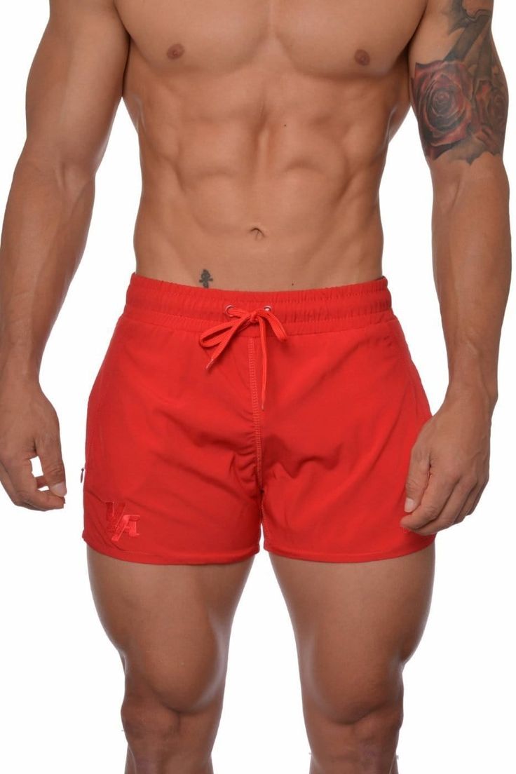 a shirtless man in red swim trunks with tattoos on his arm and chest, looking at the camera