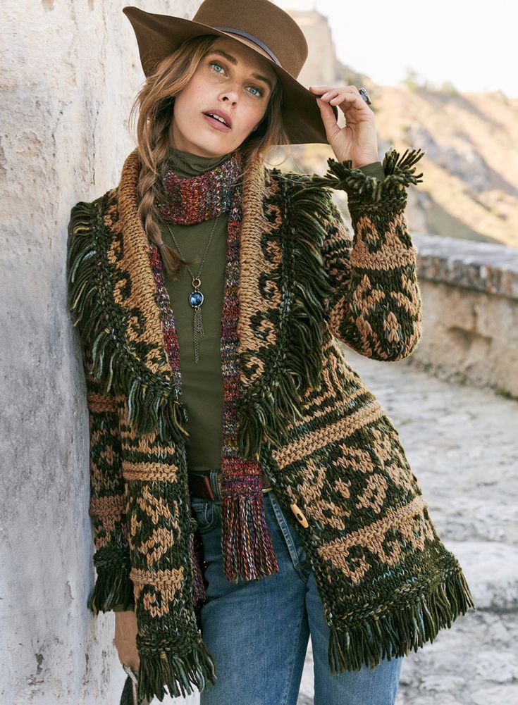 a woman wearing a brown hat and green sweater standing next to a wall with her hand on her hip