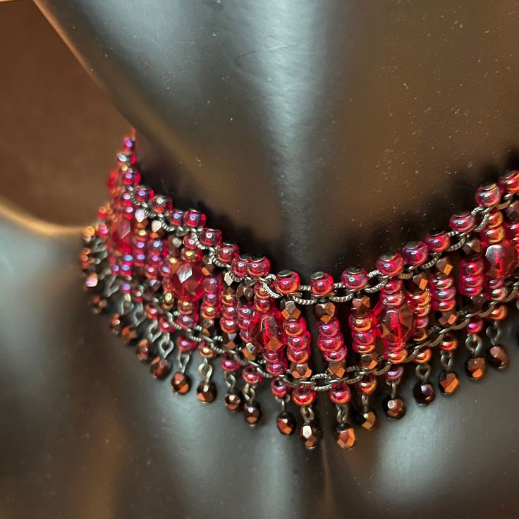 Shimmering shades of red, pink and purple in this unique, beaded collar will bring attention to the wearer. Its intricate design includes faceted red glass beads, surrounded by fat seed beads, and finished with a row of faceted, bronze-colored beads in a subtle fringe. Choker length at 15 inches, it could be extended quite nicely with a piece of grosgrain or velvet ribbon. Would look amazing with super short hair, a bob or pixie cut. I am a jewelry enthusiast, not an expert. All items are sold as described and pictured, and sourced from reputable vendors/resellers. Please, contact me with any questions. Please, refer to the images of this item as they are part of the description. Thank you. Handmade Red Beaded Bracelets For Party, Bohemian Czech Glass Beads For Party, Red Beaded Jewelry For Party, Party Choker With Dangling Round Beads, Red Beaded Bracelets With Black Beads For Festival, Party Choker With Dangling Beads, Red Beaded Chain Bracelets For Festival, Adjustable Beaded Necklace For Party, Adjustable Beaded Chain Necklace For Party