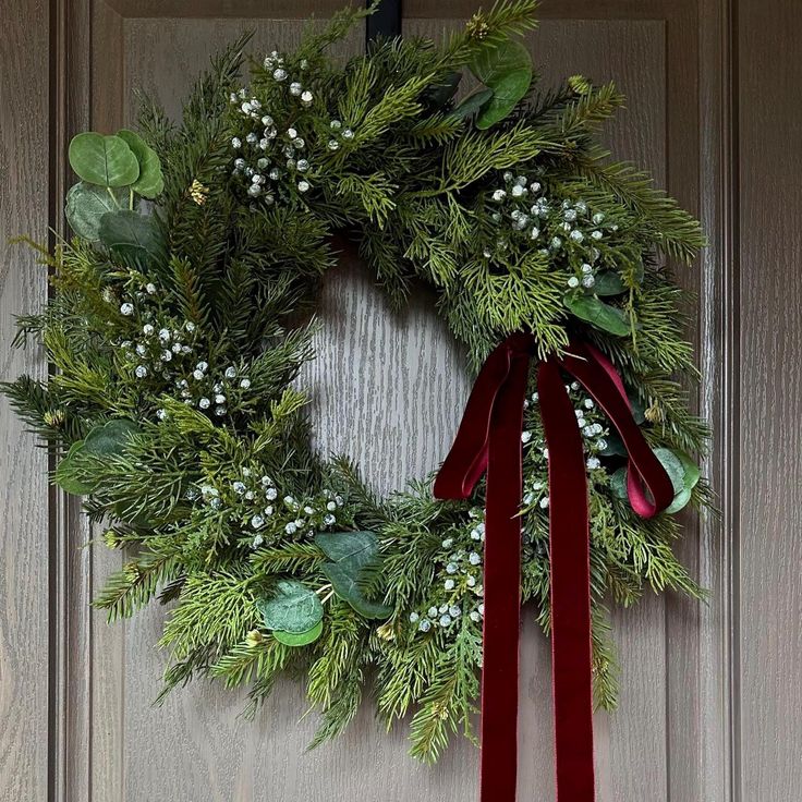 a christmas wreath hanging on a door with a red ribbon around it and greenery