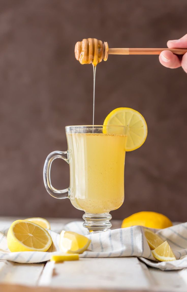 a person holding a honey stick over a cup filled with liquid and lemon wedges