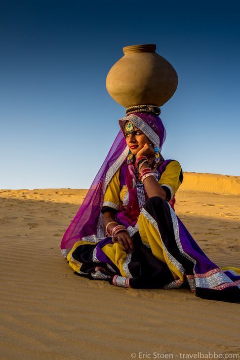 a woman sitting in the sand with a large pot on her head