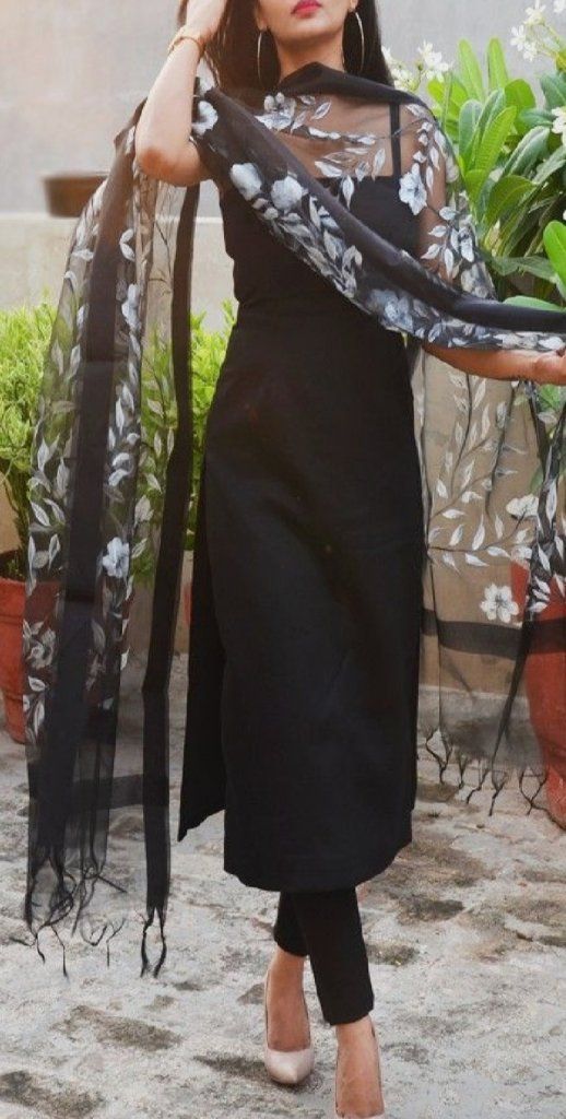 a woman wearing a black dress and scarf standing in front of potted plants with her hands on her head