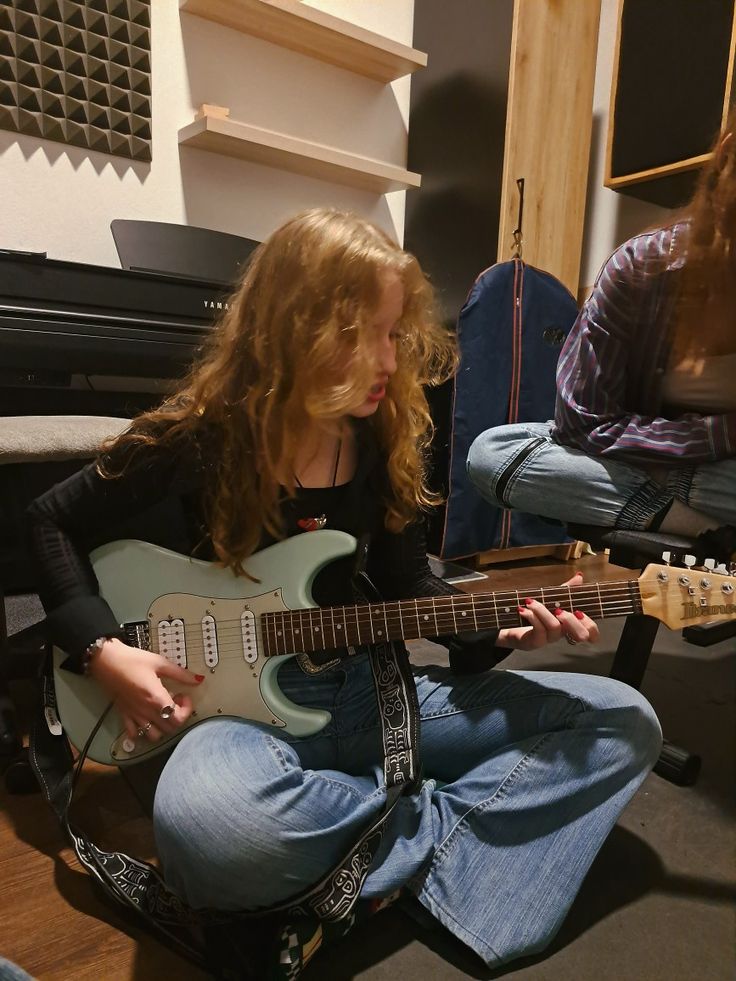 a woman sitting on the floor playing an electric guitar with another person in the background