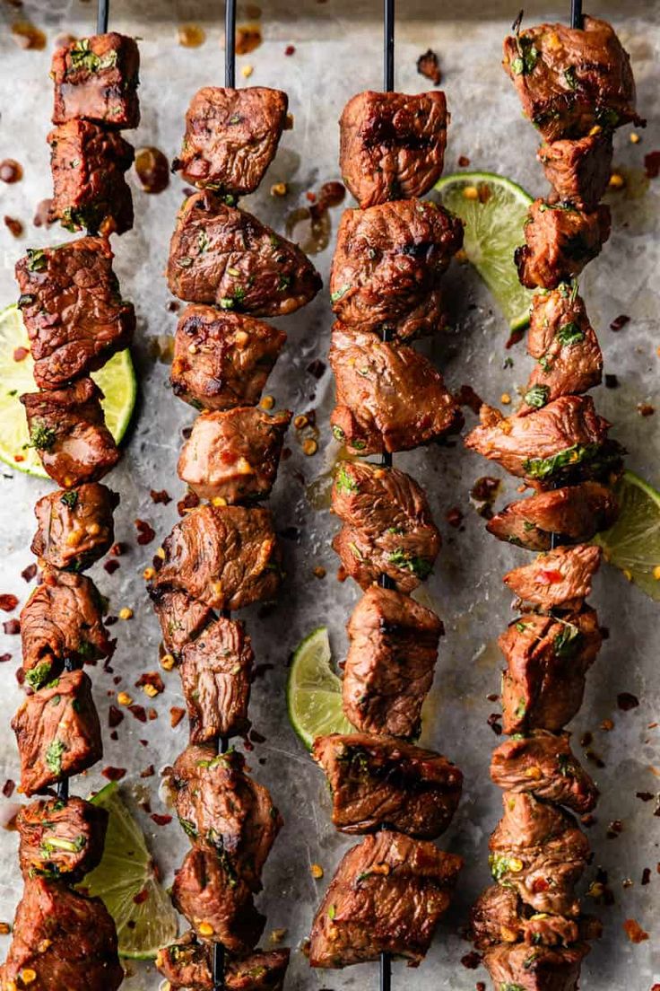 several skewers of meat with limes and seasonings on a baking sheet