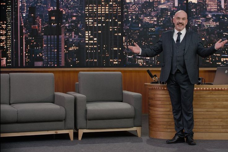 a man in a suit and tie standing next to two couches on the tonight show