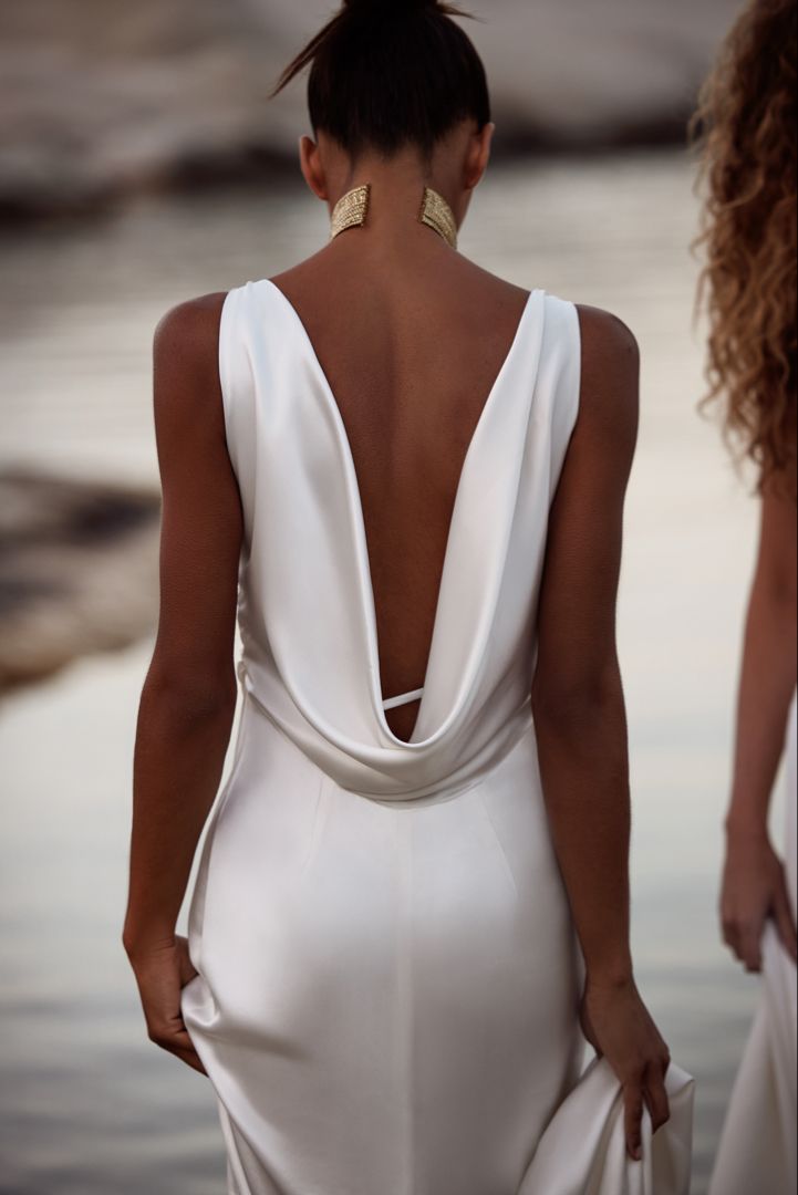 the back of a woman's white dress as she walks by water with her hands in her pockets