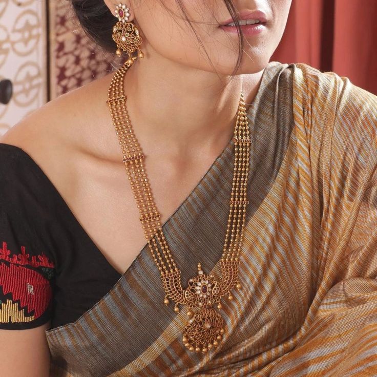 About the jewelry This long necklace set has a distinctive three-strand design and gorgeous floral motifs embedded into it. Its central attraction is a pop of color in the center. Pair it effortlessly with any saree or lehenga for a truly royal look and enchant everyone with just one look. Make it custom Want to make it a custom long necklace? Sure! Reach out to us at support@tarinika.com and we’ll be happy to make possible modifications at no extra cost. Feel free to write to us on the same ema Perhiasan India, Gold Necklace Indian, Necklace Set Indian, Gold Necklace Indian Bridal Jewelry, Long Pearl Necklaces, Antique Jewelry Indian, Bridal Fashion Jewelry, Indian Jewelry Sets, Pearl Jewelry Necklace