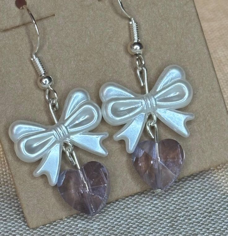 Delicate Bow and Heart Earrings These charming handmade earrings feature a pearlescent white bow charm paired with a translucent purple or pink heart-shaped bead. The bows have a lovely sheen and dimensional appearance, while the faceted heart beads add a touch of sparkle. Details: Sterling silver fish hook earwires for easy wearing Pearlescent white plastic bow charms, approximately 2 cm wide Translucent purple or pink faceted heart beads, approximately 1 cm Total drop length of about 4 cm Perfect for adding a sweet, feminine touch to any outfit. These versatile earrings would make a lovely gift or a special treat for yourself. The soft colors and dainty design make them suitable for both casual and dressier occasions. Each pair is handcrafted with care. Due to the handmade nature, slight Party Jewelry With Pink Bow In White, White Jewelry With Pink Bow For Party, White Party Jewelry With Pink Bow, Elegant Bow Heart Earrings For Party, Heart-shaped Earrings With Bow For Gift, Heart-shaped Bow Earrings For Party, Heart Shaped Bow Earrings For Party, Party Heart Earrings With Bow, White Feminine Heart Earrings For Valentine's Day