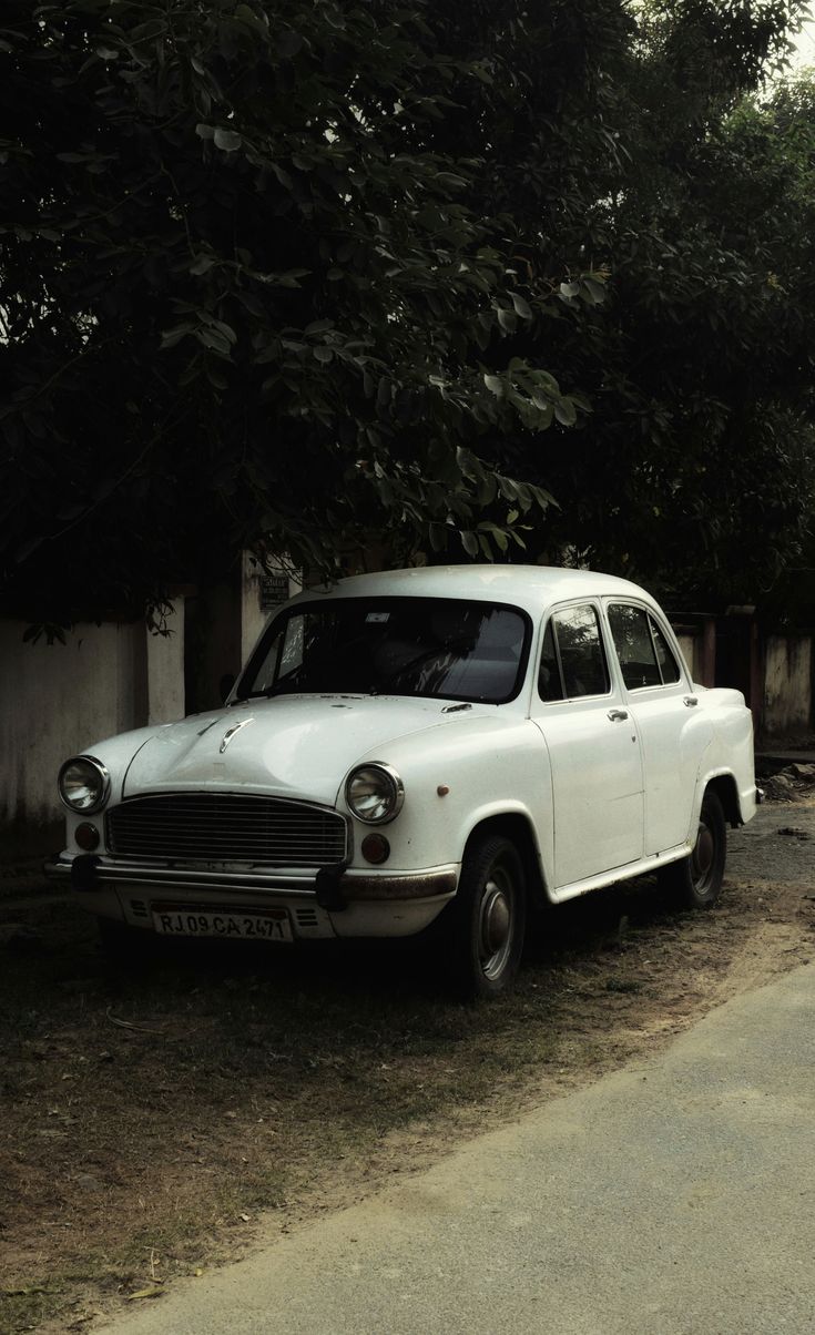 an old white car is parked on the side of the road next to a tree