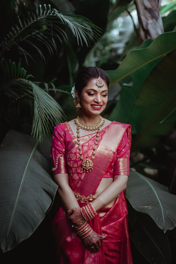 a woman in a red and gold saree standing next to some plants with her hands on her hips