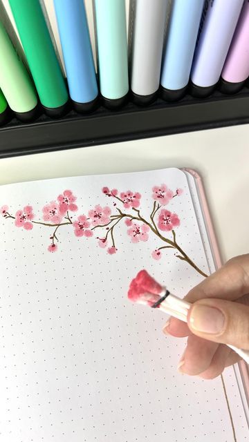 a person is holding a pen and writing on a notebook with cherry blossoms painted on it