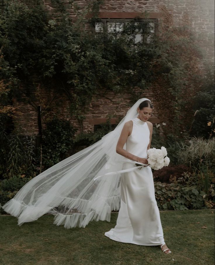 a woman in a wedding dress and veil standing on the grass with her hand over her shoulder