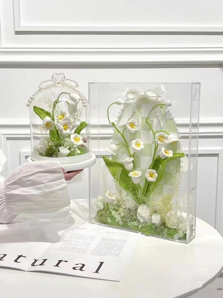 a white table topped with two vases filled with flowers and greenery next to a book