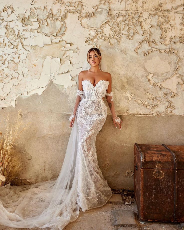 a woman standing next to a wall wearing a white dress and holding an old trunk