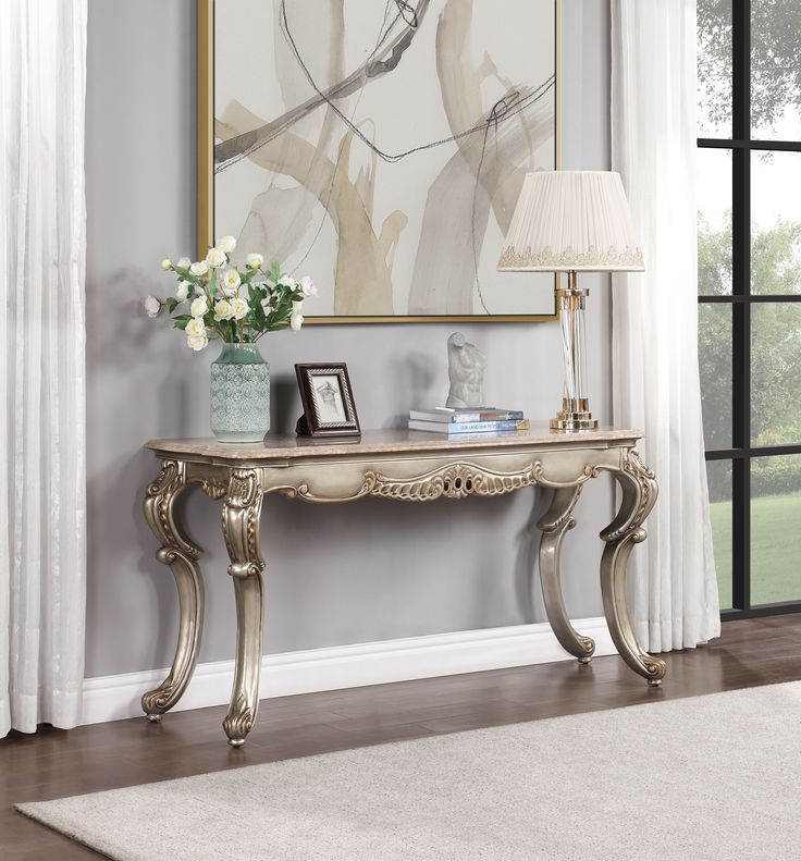 an elegant console table with flowers on it in front of a large painting and white curtains