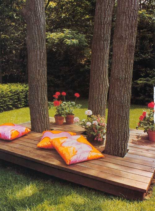 a wooden deck with pillows on it in the middle of some trees and flowers outside