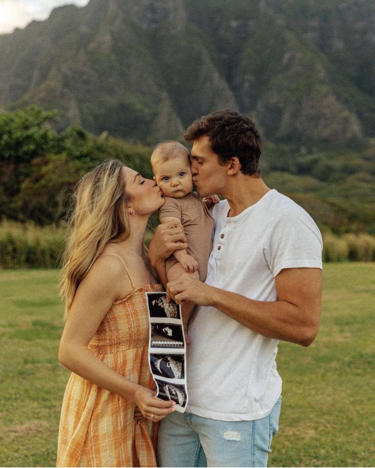 a man and woman holding a baby while standing in a field with mountains behind them