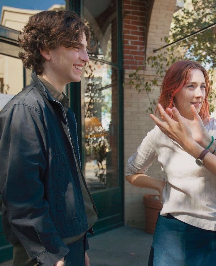 two young people standing next to each other in front of a brick building and talking