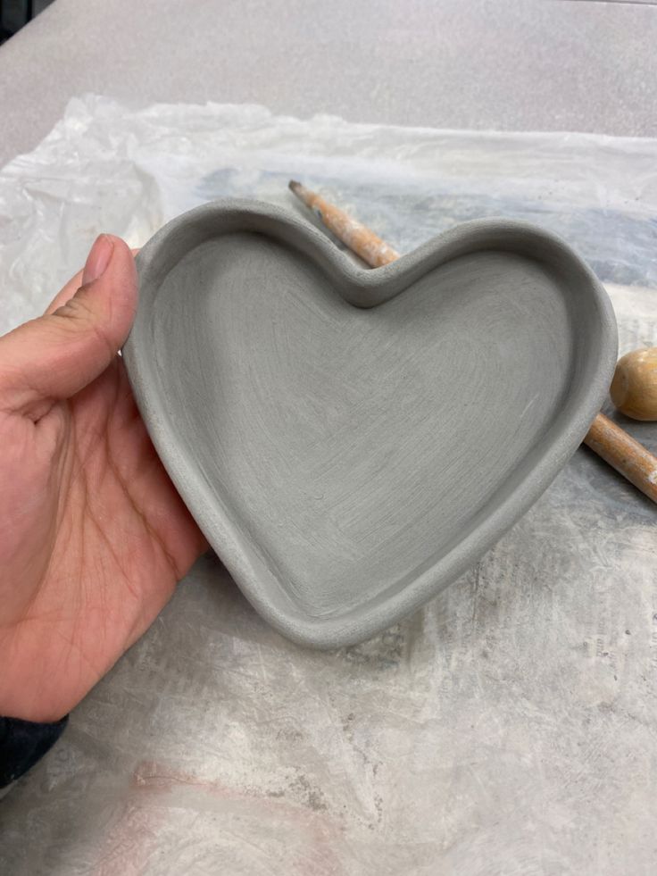 a hand holding a gray heart shaped dish with two sticks sticking out of the bowl