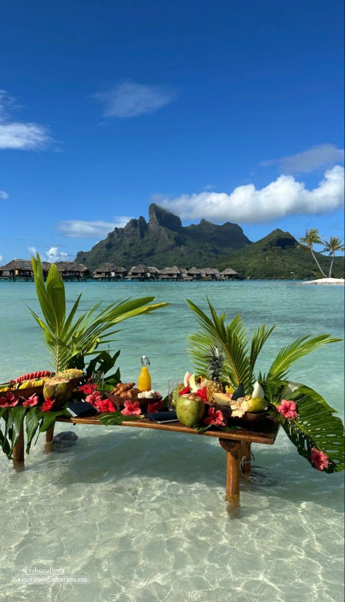 a wooden bench sitting on top of a sandy beach next to the ocean filled with flowers and fruit