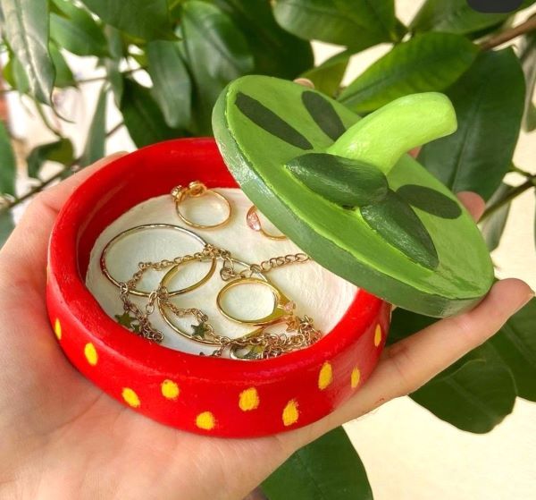 a hand holding a red ring box with gold rings in it and a green plant behind it