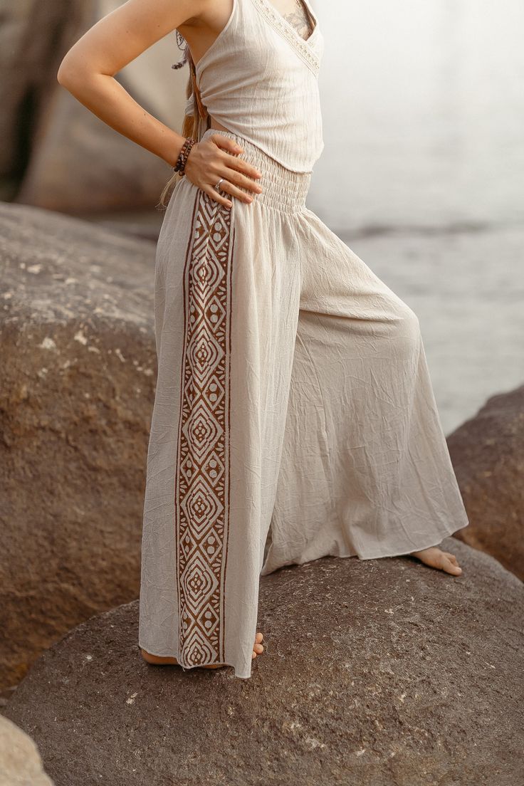 Introducing the Ragini Pants, a harmonious blend of comfort, style, and cultural charm. These wide-leg pants are crafted from soft cotton fabric, ensuring a lightweight and breathable feel that keeps you cool and relaxed throughout the day. In a serene natural color, they embody simplicity and versatility, perfect for casual outings. Adding a touch of traditional artistry, the Ragini Pants feature intricate block prints delicately adorning the sides. These block prints not only enhance the pants' aesthetic appeal but also celebrate the beauty of handcrafted designs rooted in cultural heritage. Each detail reflects a commitment to craftsmanship and timeless elegance. Designed for effortless elegance and everyday wear, the Ragini Pants offer a flattering silhouette with their wide-leg, provi Boho Print Hippie Pants For Vacation, Hippie Boho Print Pants For Vacation, Ankle-length Wide Leg Pants For Beach, Vacation Hippie Pants With Boho Print, Vacation Boho Print Hippie Pants, Bohemian Boho Print Pants For Vacation, Bohemian Wide Leg Cotton Yoga Pants, Bohemian Cotton Wide Leg Yoga Pants, Bohemian Cotton Wide Leg Ankle-length Pants