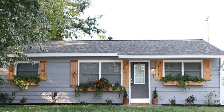 a gray house with wooden shutters and plants on the windows, in front of a tree