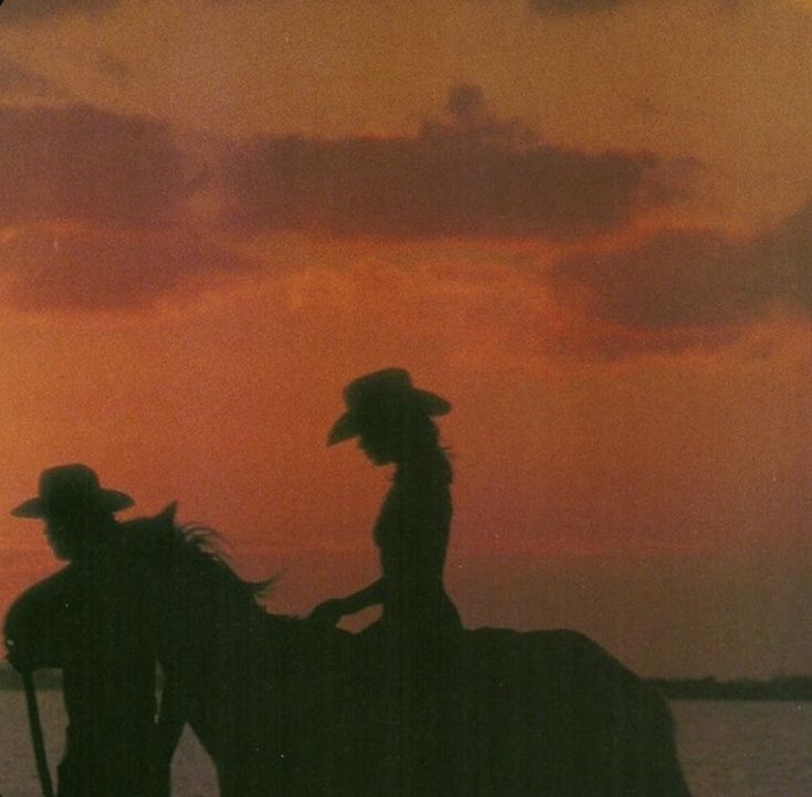 two people riding horses in front of a sunset over the ocean with clouds above them