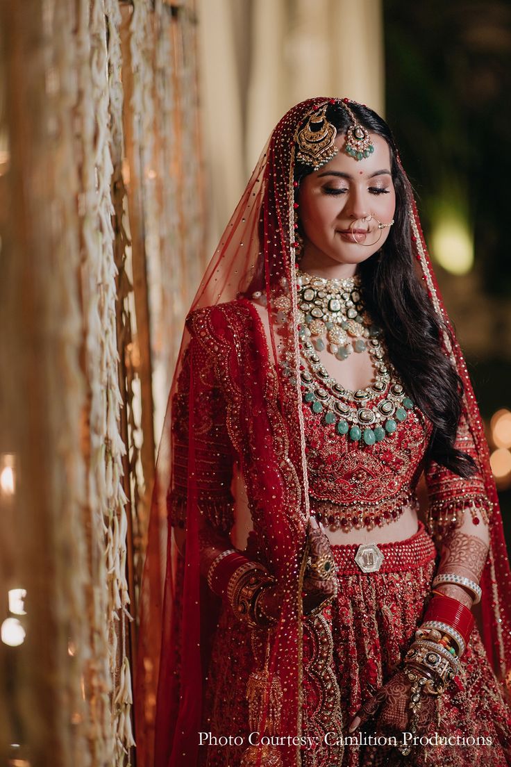 a woman in a red bridal gown and jewelry standing next to a curtain with lights behind her