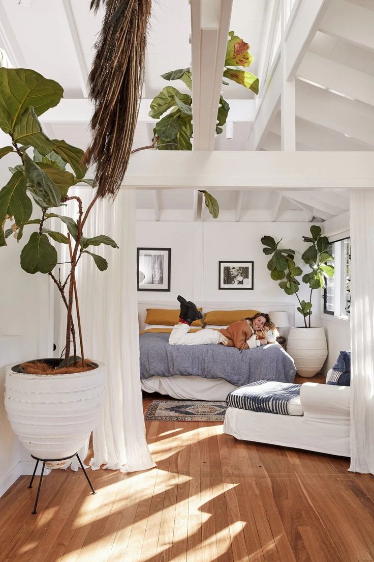 a person laying on a bed in a room with white walls and wooden floors, surrounded by potted plants