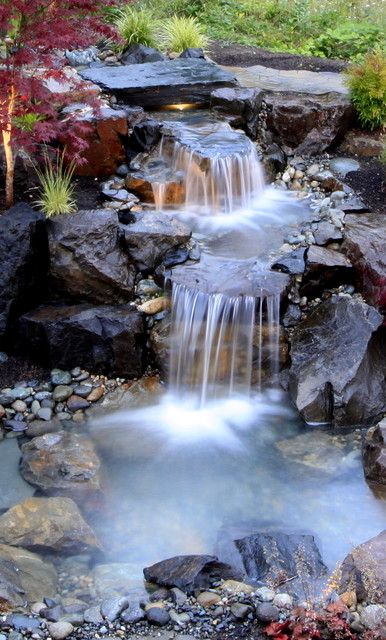 a small waterfall in the middle of a pond surrounded by rocks and trees with red leaves