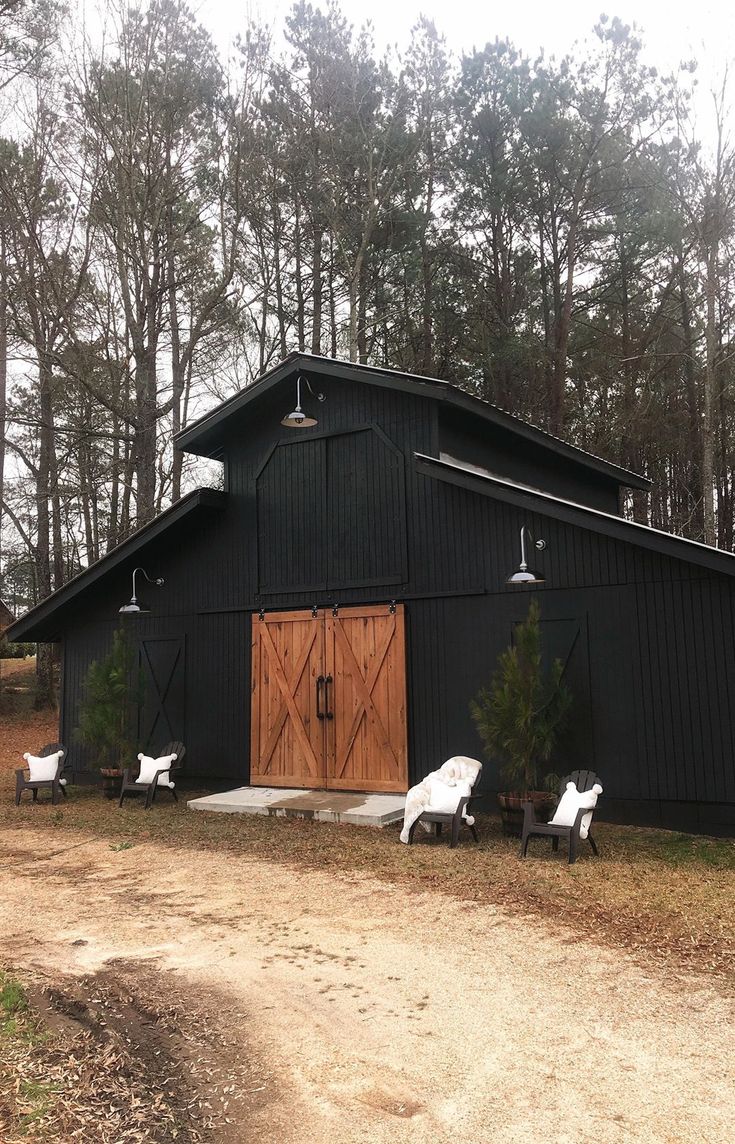 a black barn with white lawn chairs in front of it and trees around the building